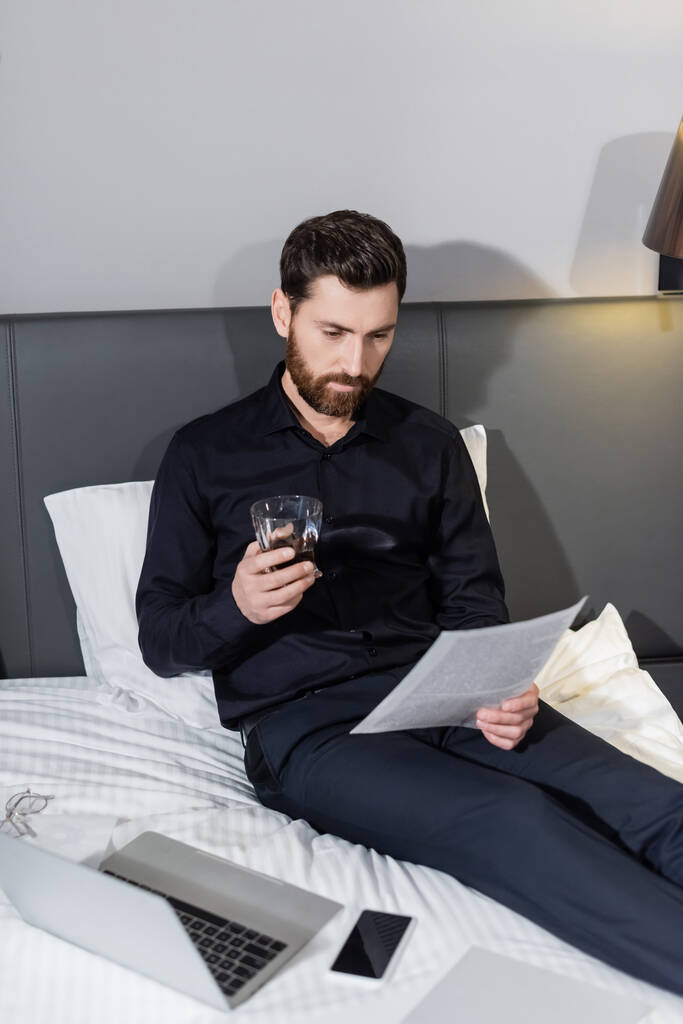 бородатый мужчина держит стакан виски и смотрит на документ в отеле - Фото, изображение