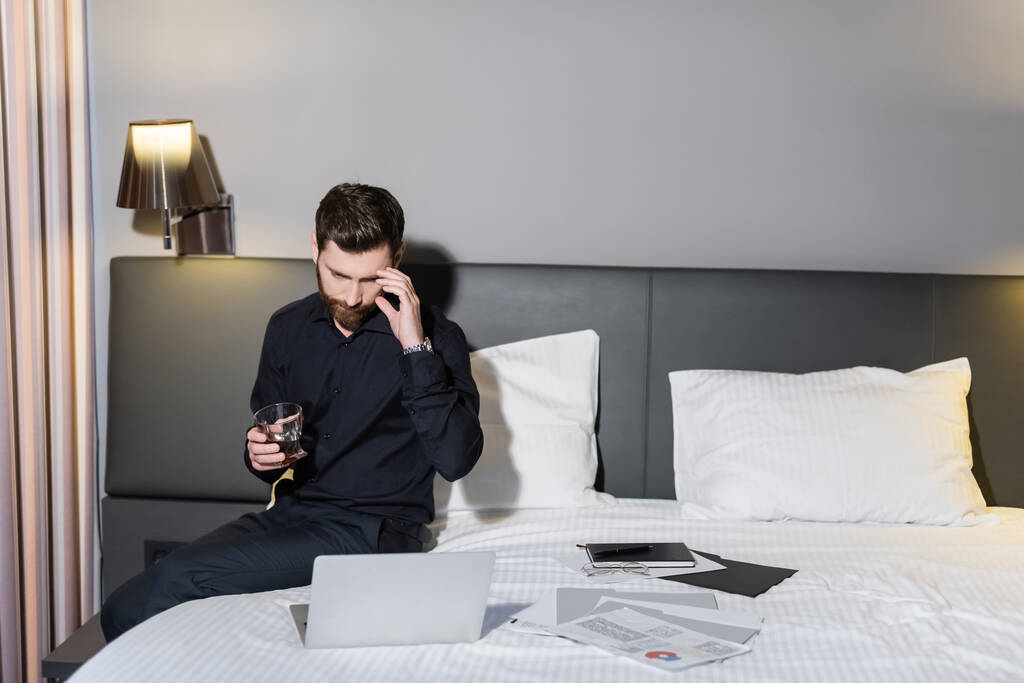 uomo barbuto in possesso di un bicchiere di whisky e guardando il computer portatile in hotel - Foto, immagini