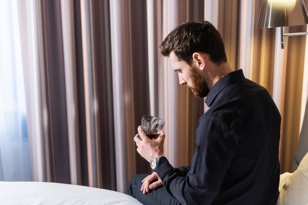 barbudo sosteniendo un vaso de whisky y sentado en la cama del hotel - Foto, imagen