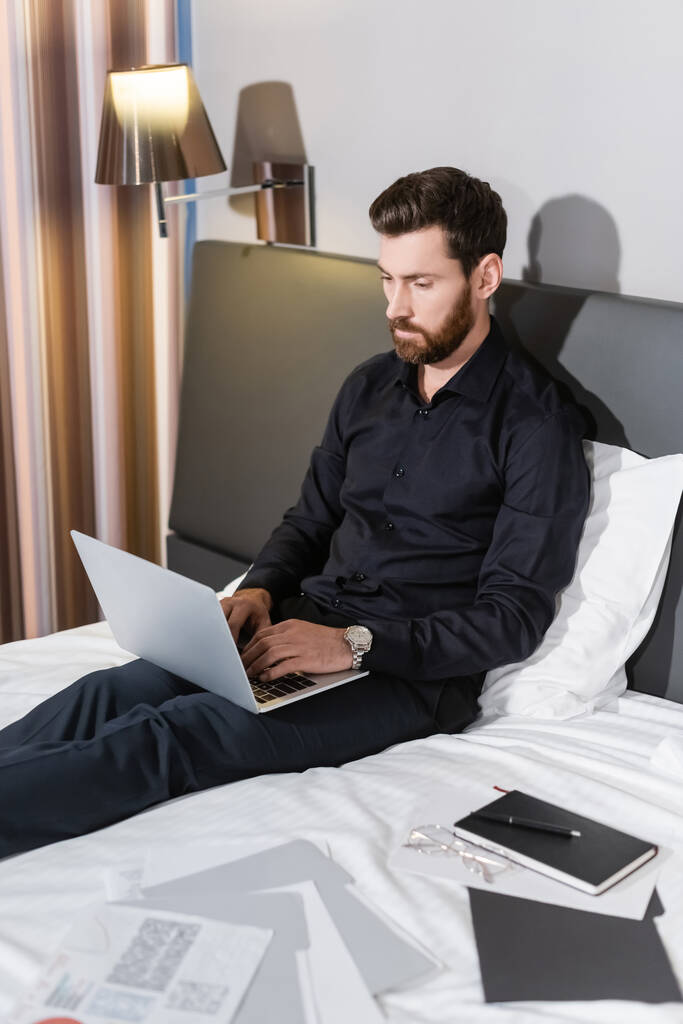 髭を生やした男がノートパソコンを使ってホテルのベッドに座り - 写真・画像