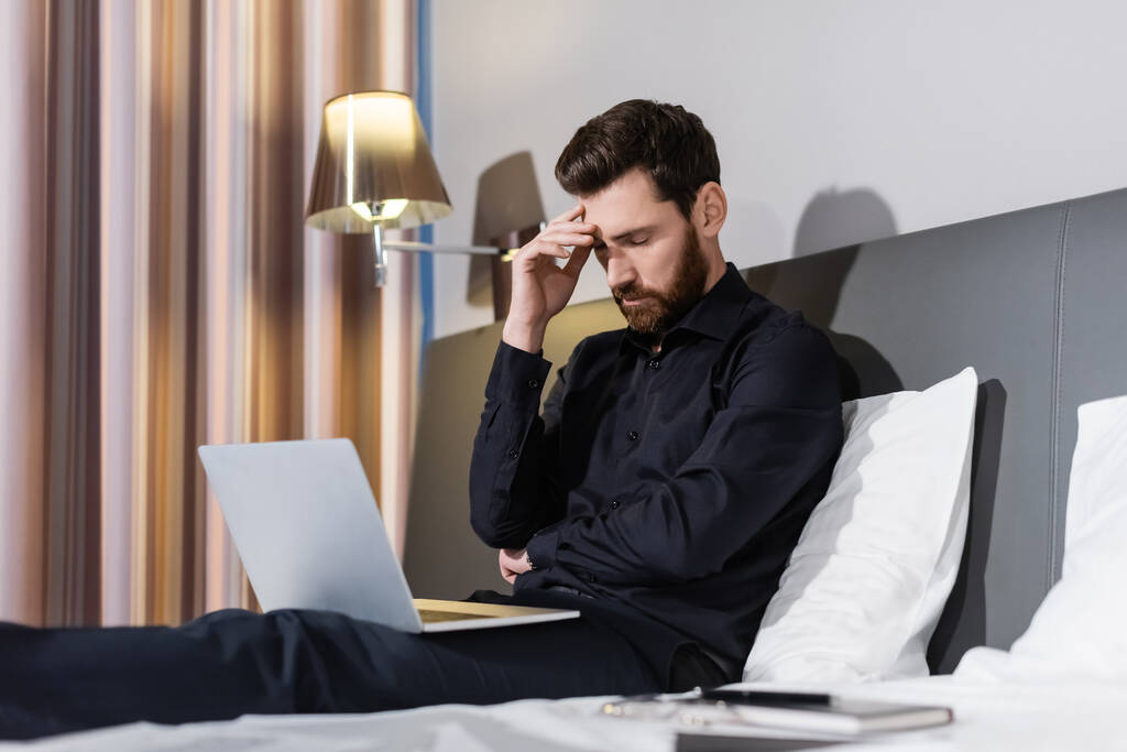 hombre barbudo cansado sentado con portátil en la cama del hotel - Foto, Imagen