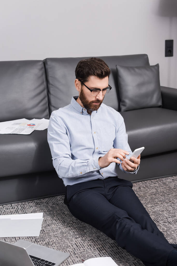 szakállas szabadúszó szemüveg ül a szőnyegen, és használja okostelefon közelében laptop és kanapé  - Fotó, kép