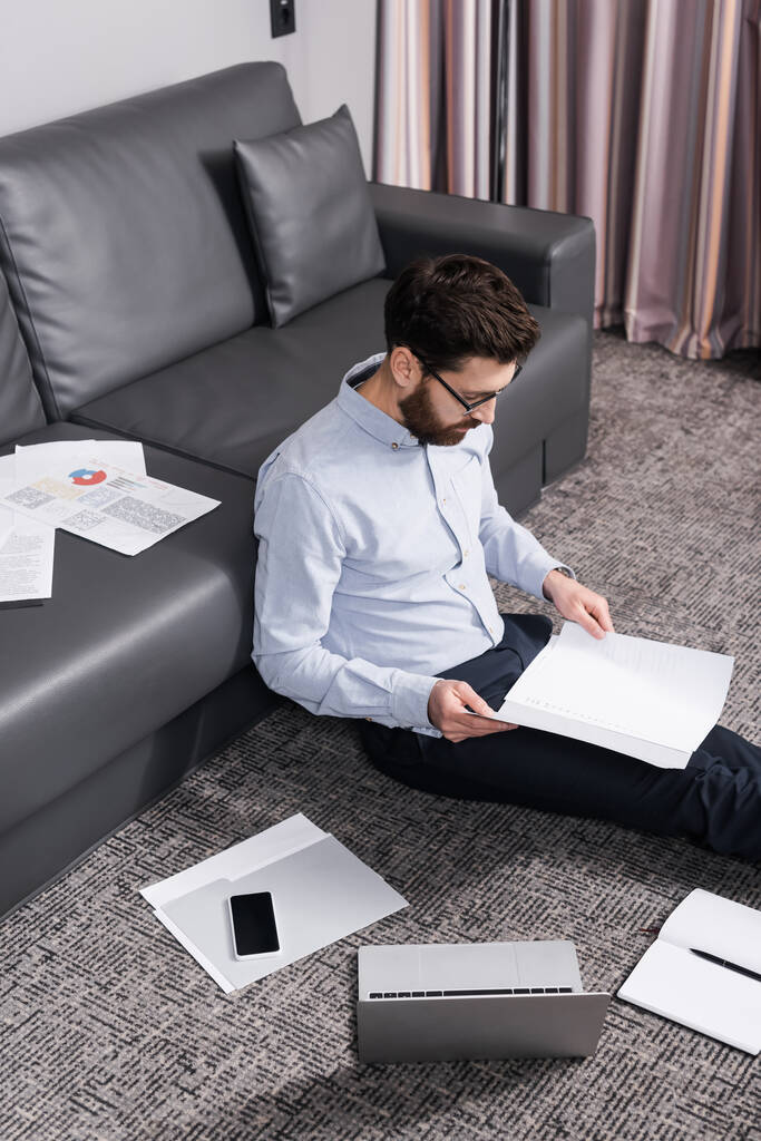 high angle view of bearded freelancer in eyeglasses sitting on carpet near documents and gadgets  - Photo, Image