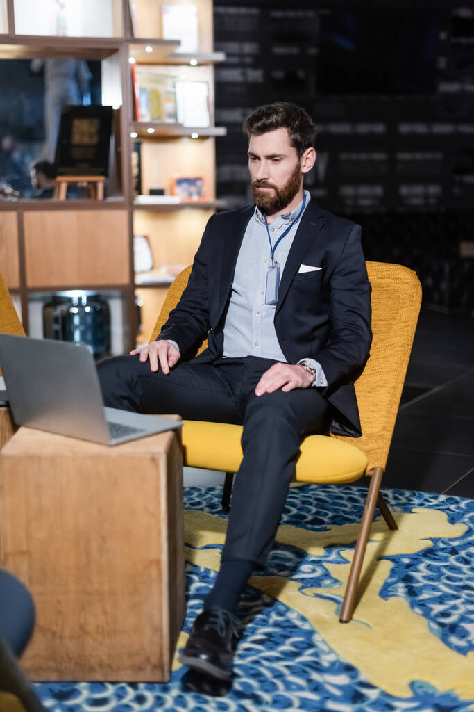 hombre barbudo con placa de identificación mirando a la computadora portátil mientras está sentado en el vestíbulo del hotel  - Foto, imagen