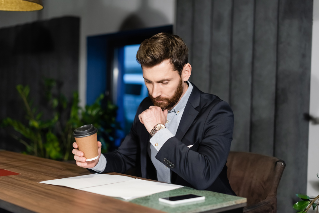 вдумчивый человек в костюме держит бумажную чашку во время просмотра документов в гостиничном кафе - Фото, изображение