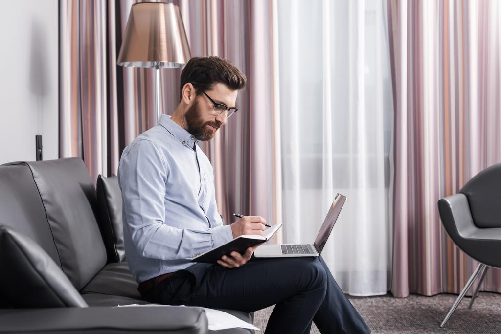 man in shirt and eyeglasses writing on notebook near laptop in hotel room  - Photo, Image
