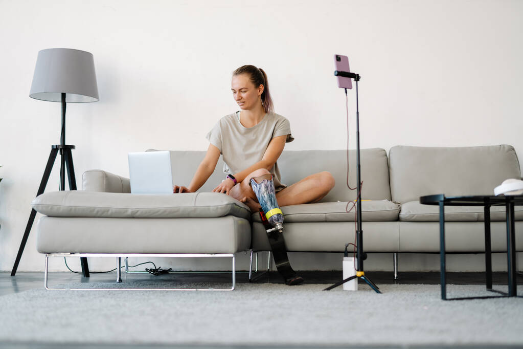 Gülümseyen beyaz engelli kadın blogcu protez bacaklı bir kadın kanepenin önünde oturmuş evde video çekiyor. - Fotoğraf, Görsel