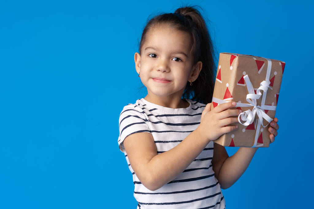 Glückliches kleines Mädchen mit langen Haaren hält eine Geschenkschachtel auf blauem Hintergrund - Foto, Bild