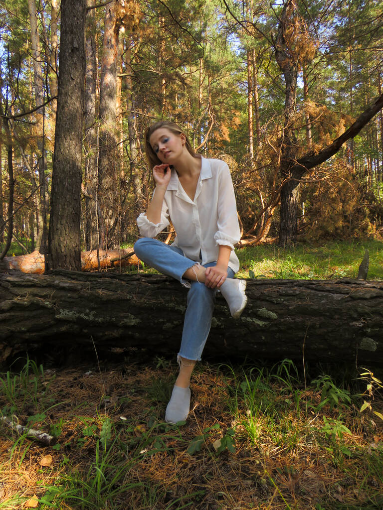 Mladá blondýna v bílé košili a modré džíny v podzimním lese na spadlém kmeni stromu se těší dovolenou v přírodě, povaha zabadnaya Sibiře, koncept outdoorových aktivit, Turistika v lese, cestování a cestovní ruch - Fotografie, Obrázek