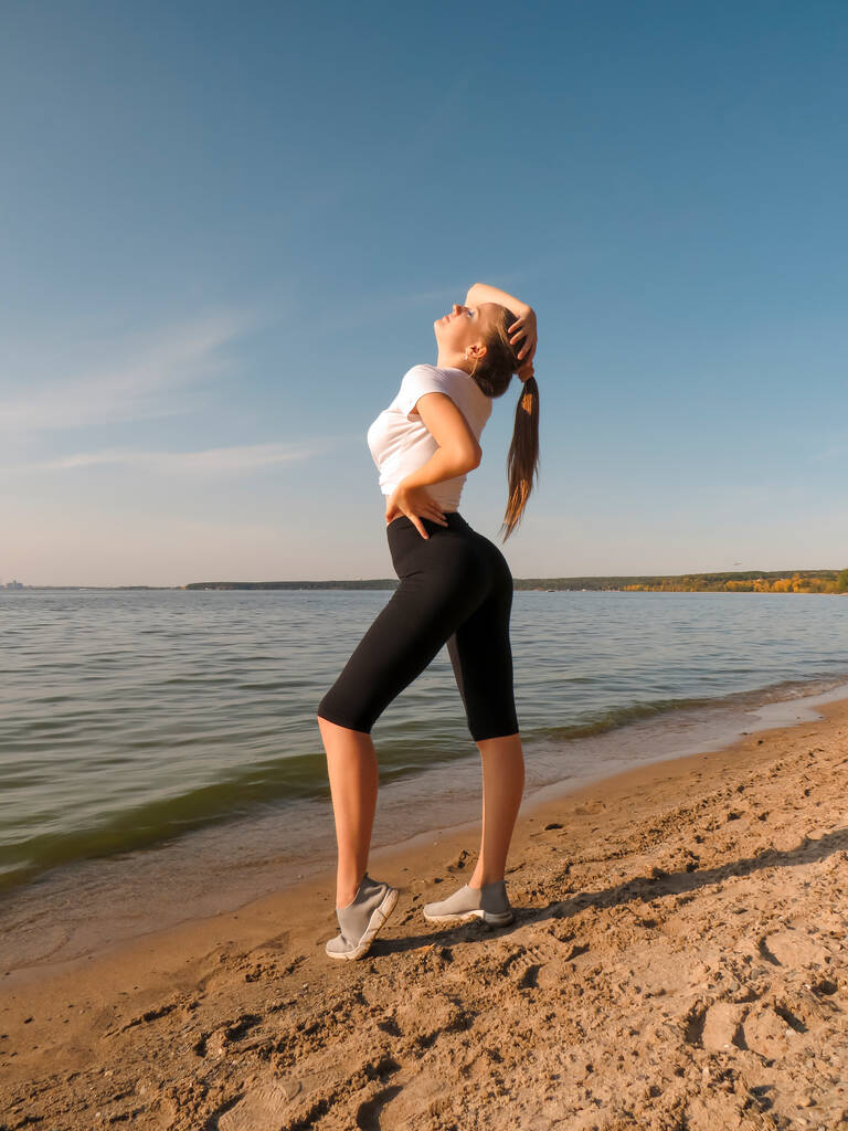 Una giovane donna bionda snella con una figura sexy in pantaloncini sportivi e una maglietta fa yoga all'aria aperta sulla riva del mare in una giornata di sole tiepido.Concetto di sport all'aria aperta, stile di vita sano, attività all'aria aperta, - Foto, immagini