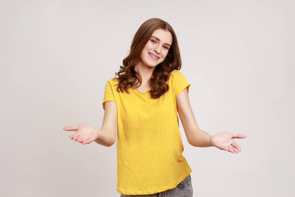Ti prego, prendine gratis! Amichevole bella giovane donna in giallo T-shirt accogliente a braccia aperte e sorridente gentilmente, guardando sorridente alla macchina fotografica. Indoor studio girato isolato su sfondo grigio. - Foto, immagini