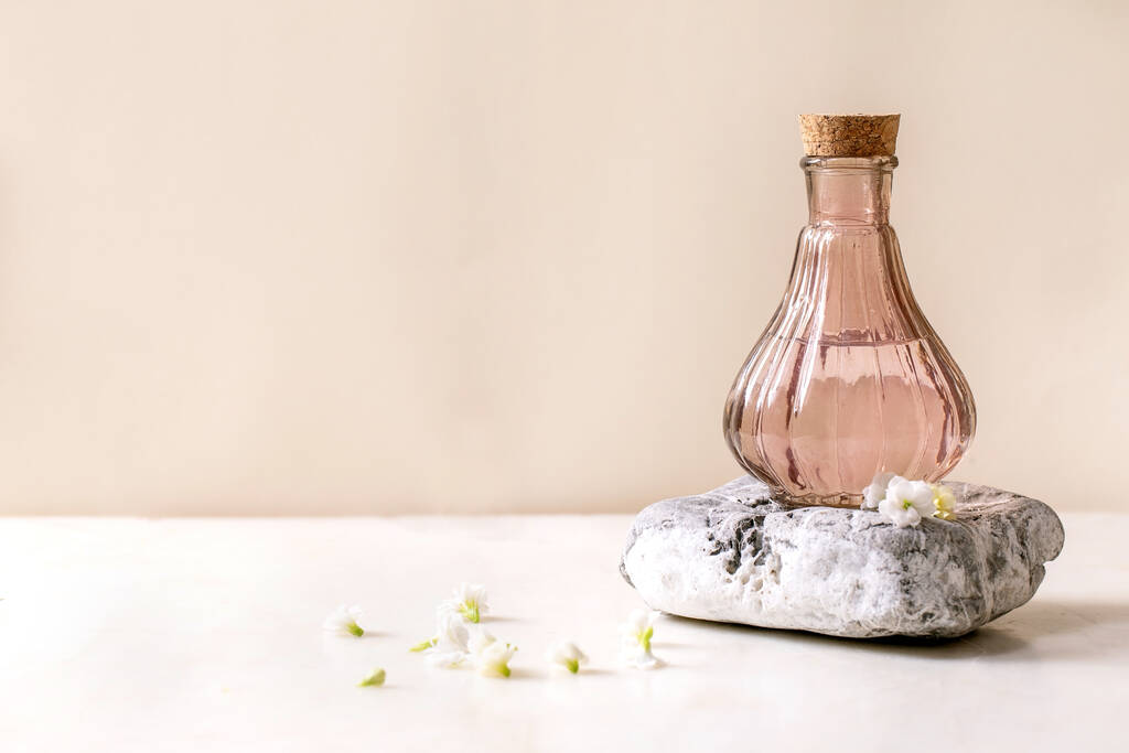 Schöne rosa transparente Flasche Parfüm mit Kork, steht auf strukturiertem Stein auf weißem Marmor Tisch mit kleinen weißen Blumen Kalanchoe herum. Parfümeriekonzept mit Platz für Design-Etikett - Foto, Bild