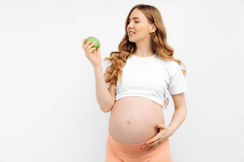 Gezonde zwangere vrouw eet een groene appel rijk aan vitaminen op een witte achtergrond. Zwangere vrouw leidt een gezonde levensstijl, genieten van een gezond dieet tijdens de zwangerschap. Zwangerschap, gezond voedsel - Foto, afbeelding