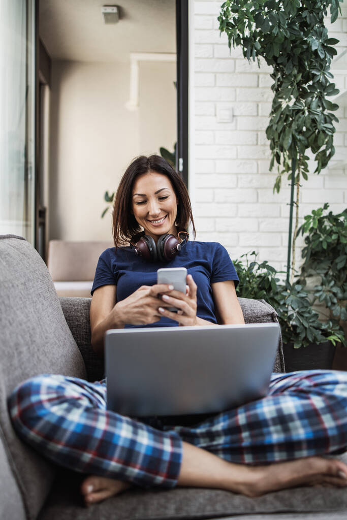 Giovane donna adulta sdraiata sul divano nel suo soggiorno e utilizzando il computer portatile per la navigazione web e la comunicazione e la chat sui social media. - Foto, immagini