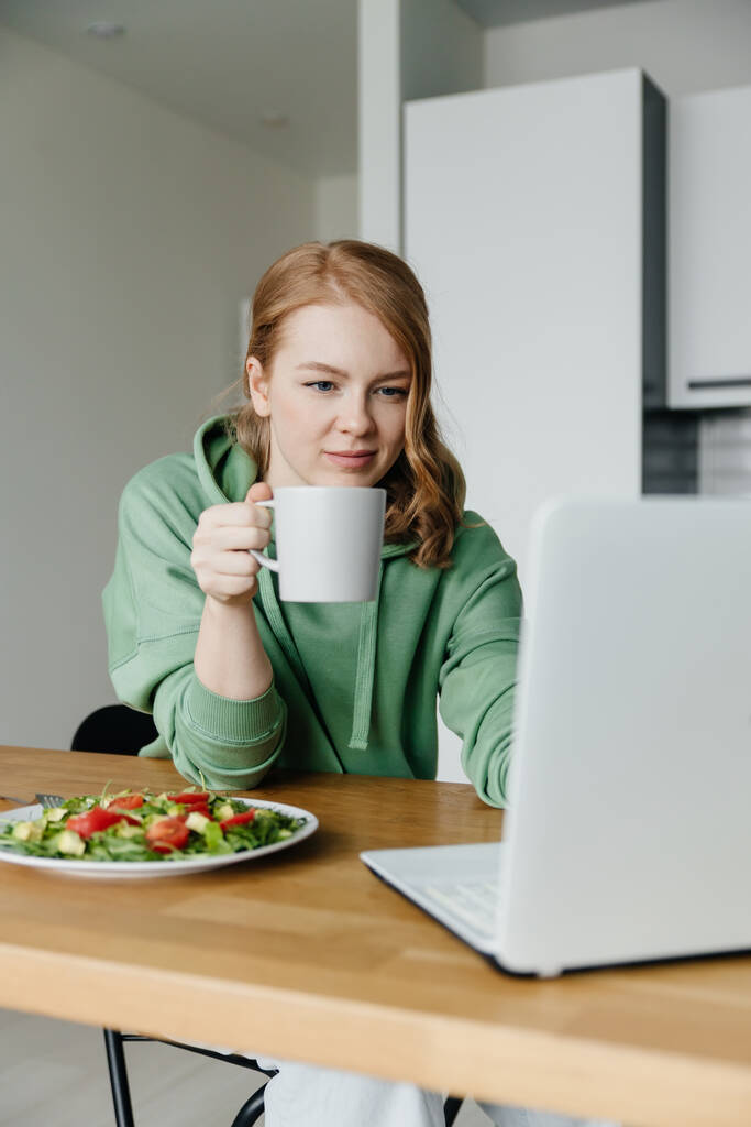 Mujer comiendo ensalada y trabajando usando laptop en su cocina - Foto, imagen