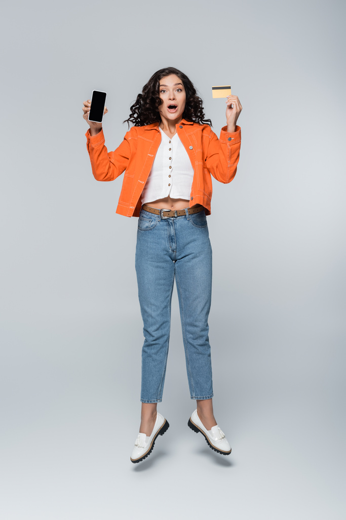 pełna długość zaszokowanej kobiety w pomarańczowej kurtce z kartą kredytową z kasą i smartfonem lewitującej na szarym - Zdjęcie, obraz