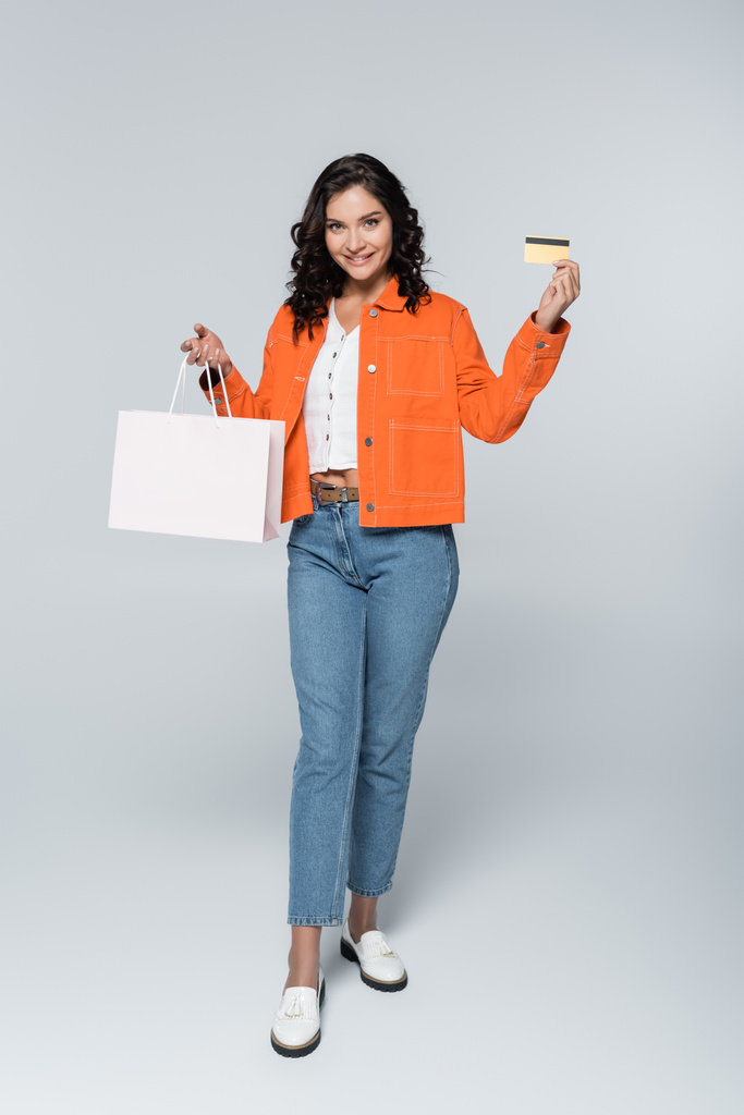 полная длина счастливой женщины в оранжевой куртке, держащей кредитную карту с возвратом денег и сумку с покупками на сером - Фото, изображение