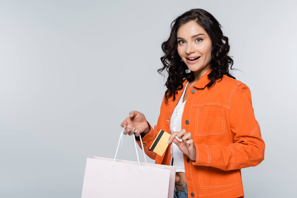 mulher animada em casaco laranja segurando cartão de crédito com cashback perto de saco de compras isolado em cinza - Foto, Imagem