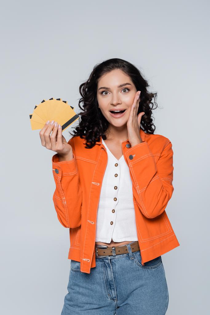 verbaasd jonge vrouw in oranje jas houden credit cards met cashback geïsoleerd op grijs - Foto, afbeelding