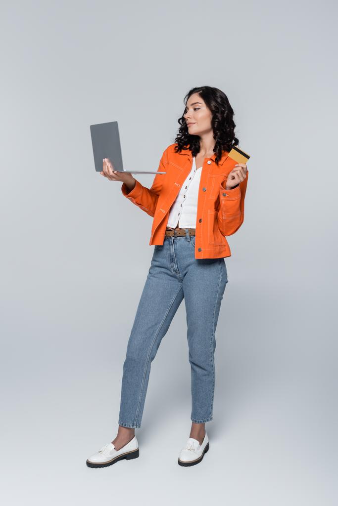 longitud completa de la mujer joven en chaqueta naranja que sostiene el ordenador portátil y la tarjeta de crédito en gris  - Foto, imagen
