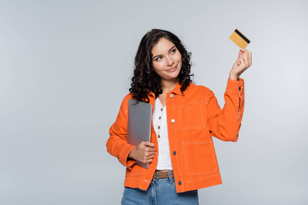 tevreden vrouw in oranje jas houden laptop en kijken naar credit card geïsoleerd op grijs  - Foto, afbeelding