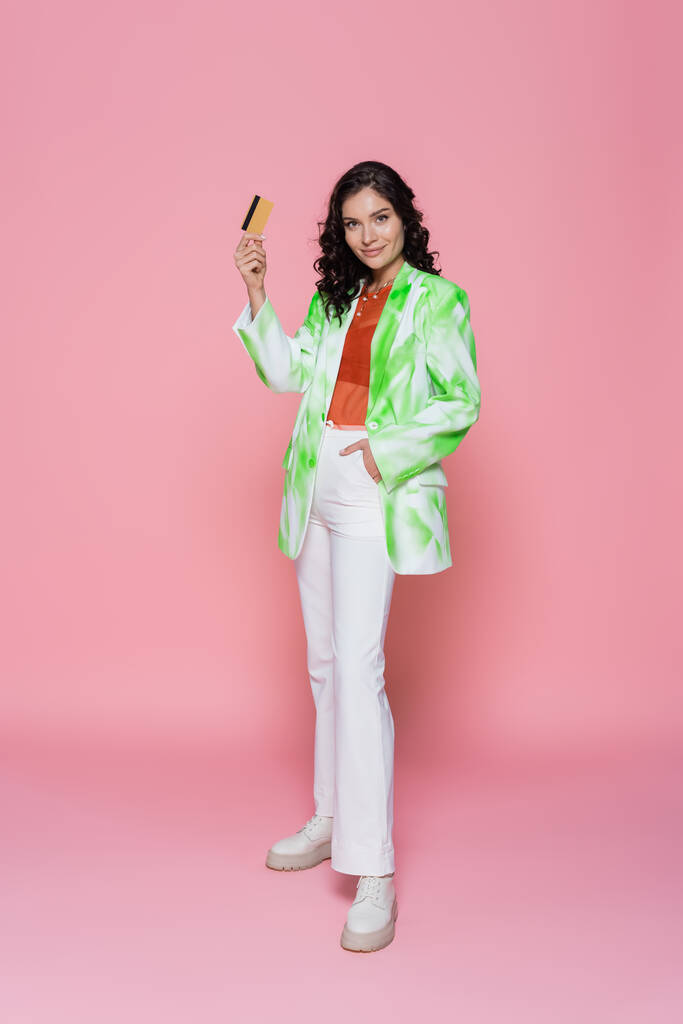 full length of pleased young woman in tie-dye blazer holding credit card and posing with hand in pocket on pink  - Photo, Image