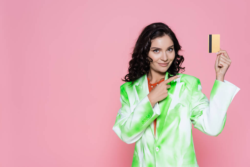 cheerful young woman in tie-dye blazer pointing at credit card isolated on pink  - Photo, Image