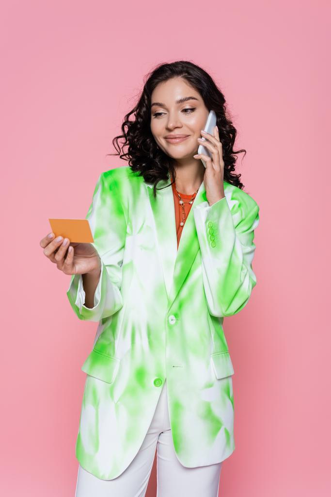 jeune femme souriante en blazer de teinture cravate tenant la carte de crédit et parlant sur smartphone isolé sur rose  - Photo, image