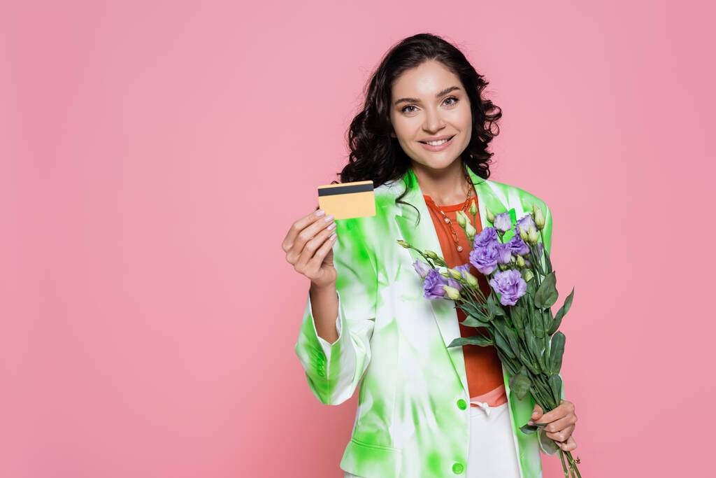 mosolygós fiatal nő nyakkendő festék blézer gazdaság hitelkártya és virágok elszigetelt rózsaszín  - Fotó, kép