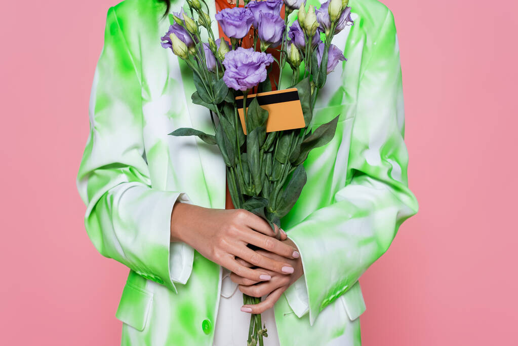 vista ritagliata di giovane donna in cravatta blazer colorante in possesso di fiori viola con carta di credito isolata su rosa  - Foto, immagini