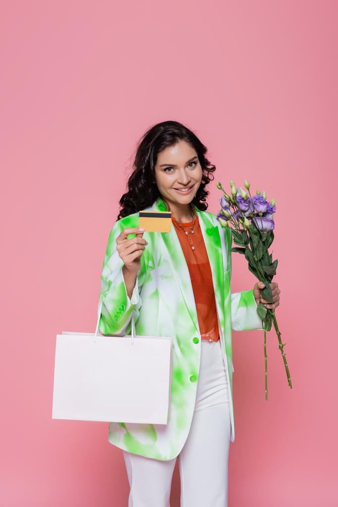 Kravat boyalı ceketli gülümseyen genç bir kadın. Elinde kredi kartı, alışveriş çantası ve pembe üzerine izole edilmiş çiçekler var.  - Fotoğraf, Görsel
