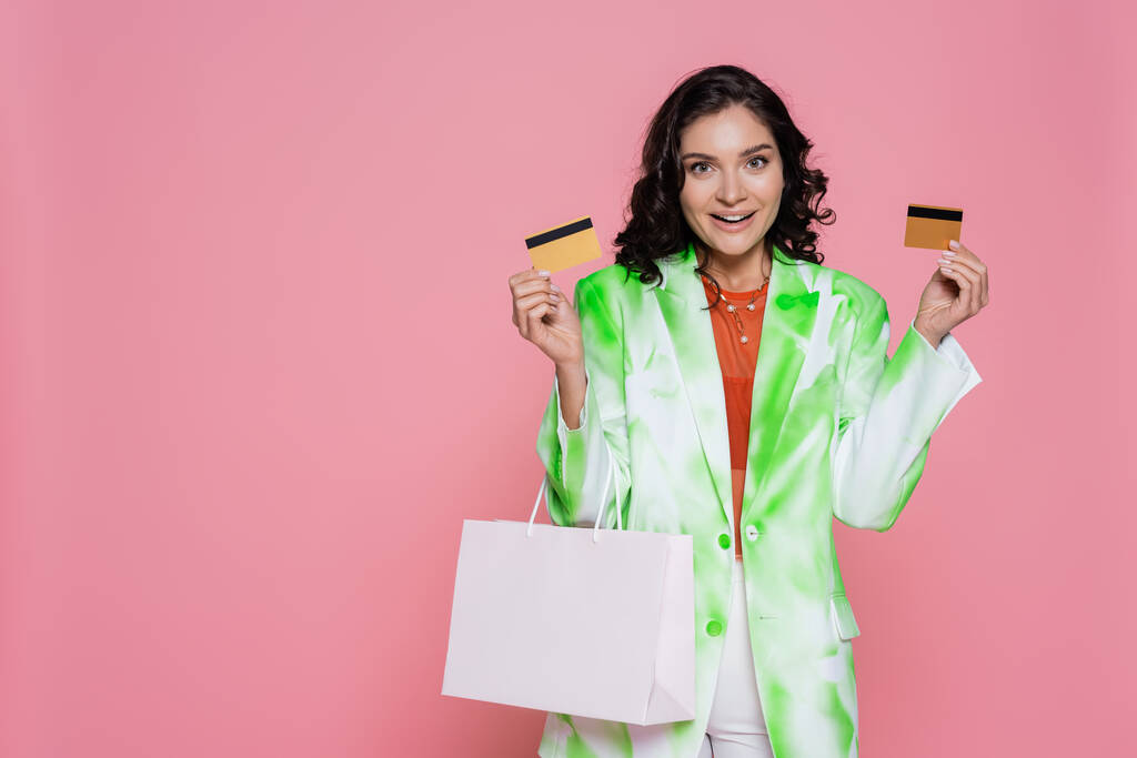 Heyecanlı genç bir kadın kravat boyası ceketli, elinde kredi kartları ve pembe bir alışveriş çantası var.  - Fotoğraf, Görsel