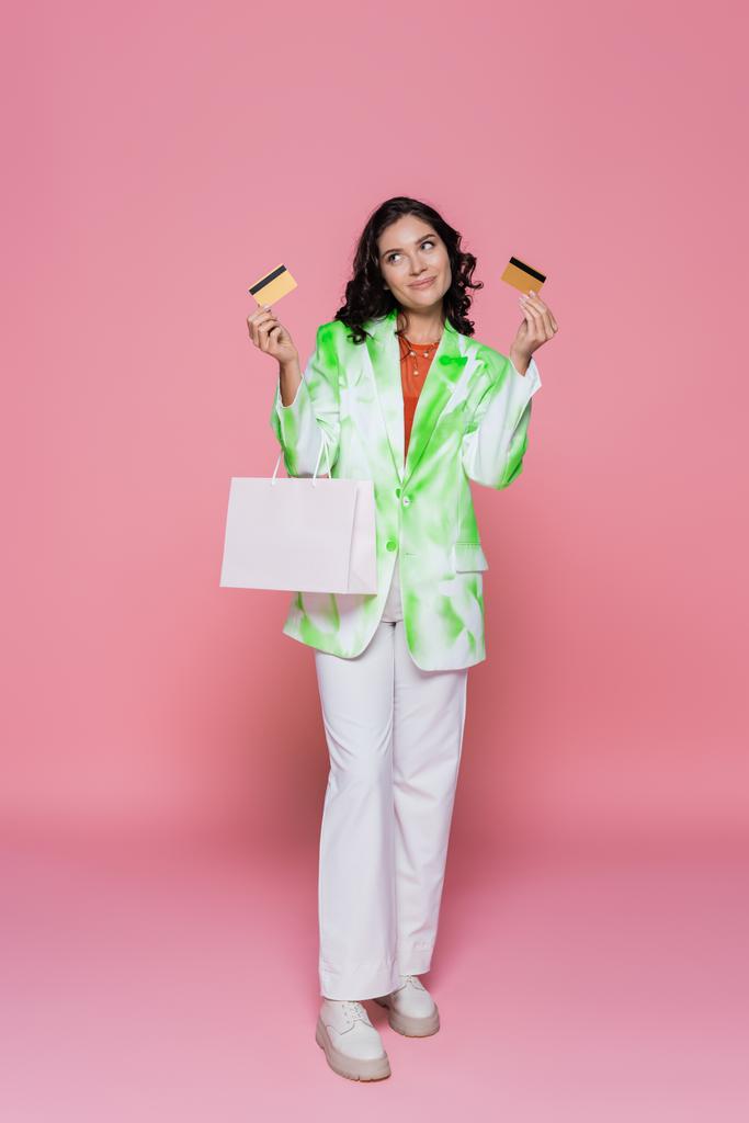 volledige lengte van de lachende vrouw in tie dye blazer met creditcards en boodschappentas op roze  - Foto, afbeelding