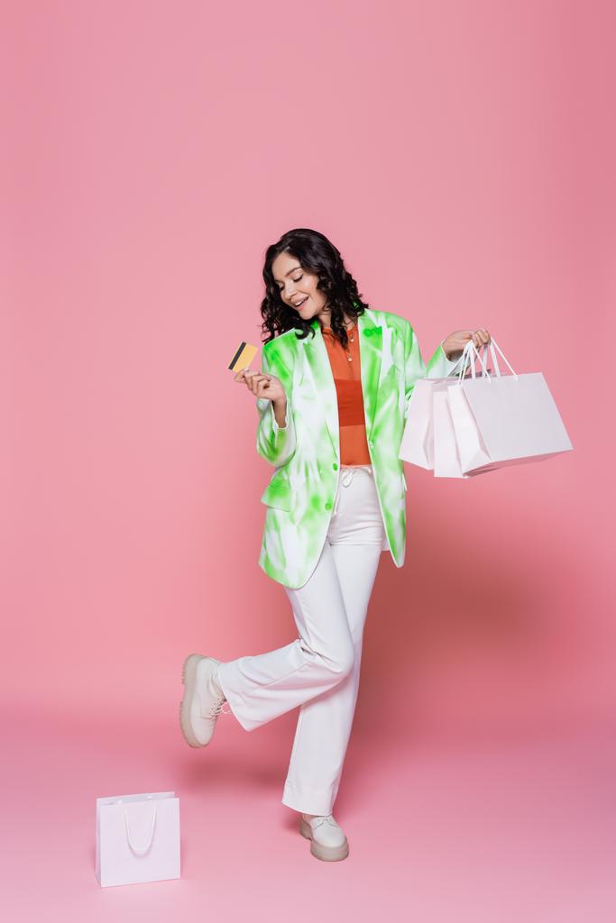 teljes hossza vidám fiatal nő nyakkendő festék blézer gazdaság hitelkártya és bevásárló táskák rózsaszín  - Fotó, kép