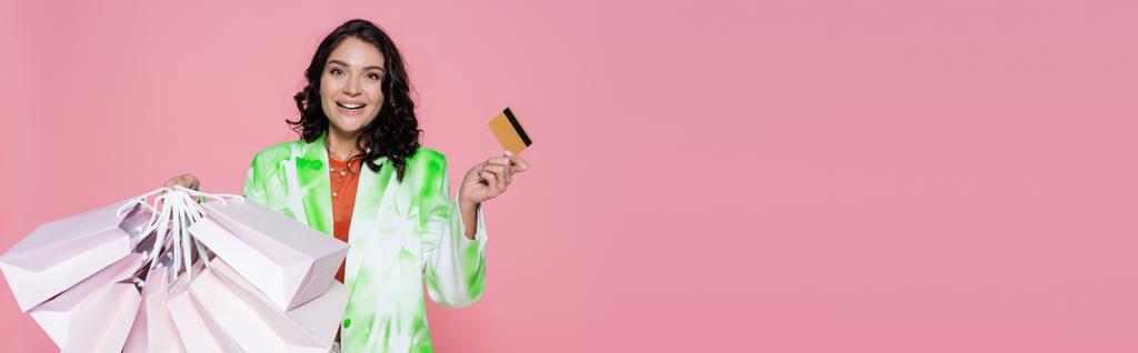 alegre joven mujer en corbata blazer tinte celebración de tarjetas de crédito y bolsas de compras aislados en rosa, bandera - Foto, Imagen