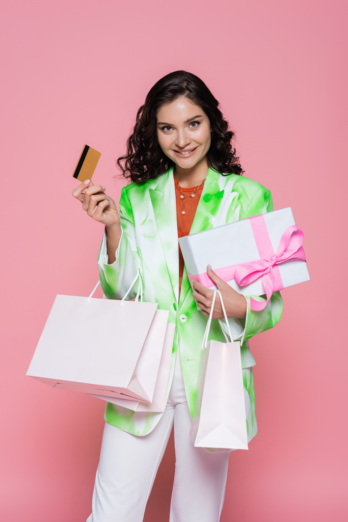 alegre joven mujer en corbata chaqueta de tinte celebración de tarjeta de crédito, bolsas de compras y regalo envuelto aislado en rosa - Foto, imagen