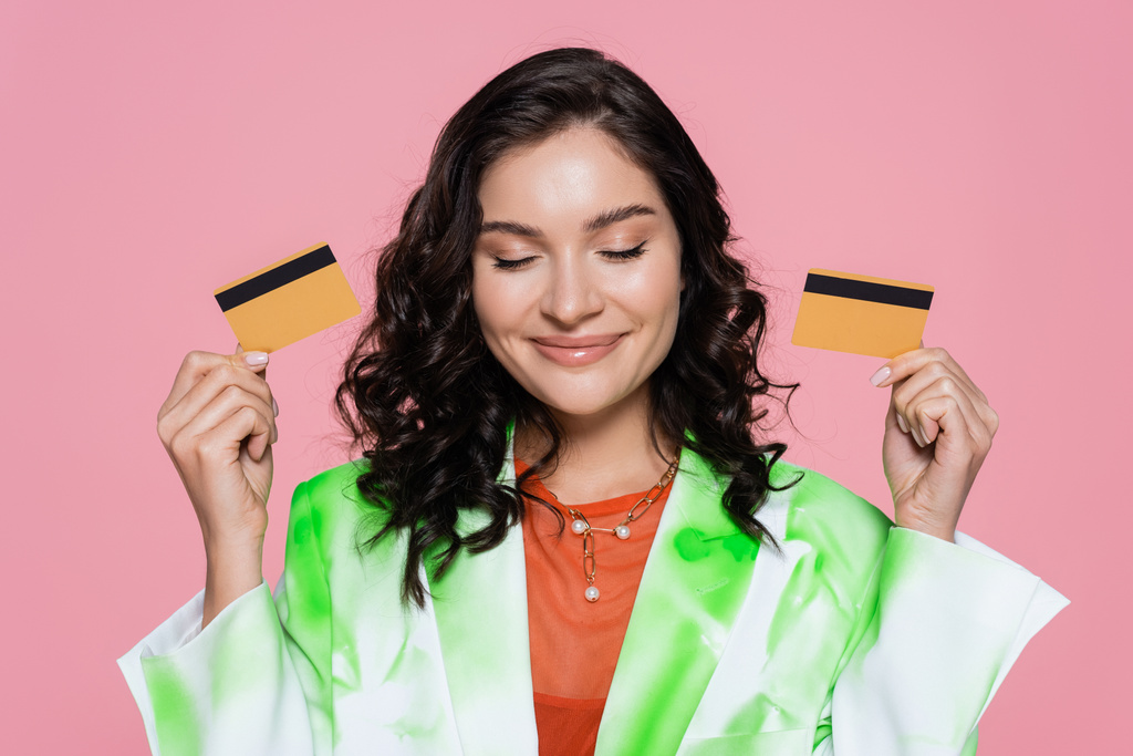 zufriedene Frau in grünem Krawattenblazer, Kreditkarten in der Hand und isoliert lächelnd auf rosa - Foto, Bild