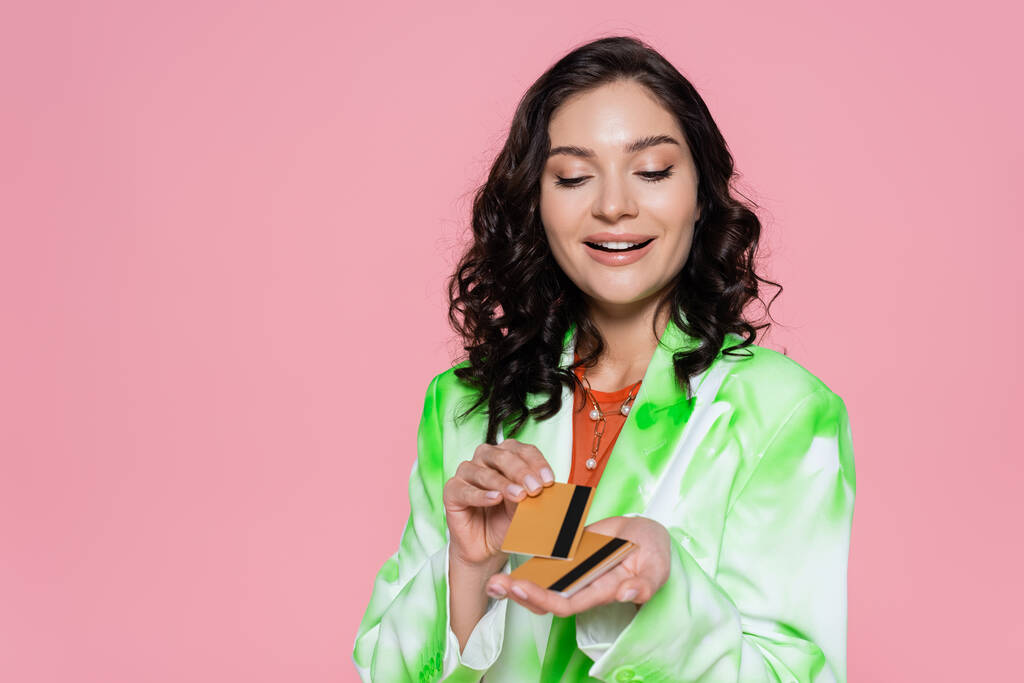 radostná žena v zelené kravatě barvivo blejzr při pohledu na kreditní karty a usmívá izolované na růžové - Fotografie, Obrázek