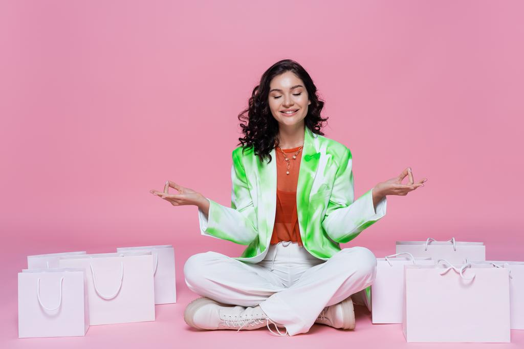 šťastná brunetka žena v blejzru sedí v józe póza při meditaci v blízkosti nákupních tašek na růžové  - Fotografie, Obrázek