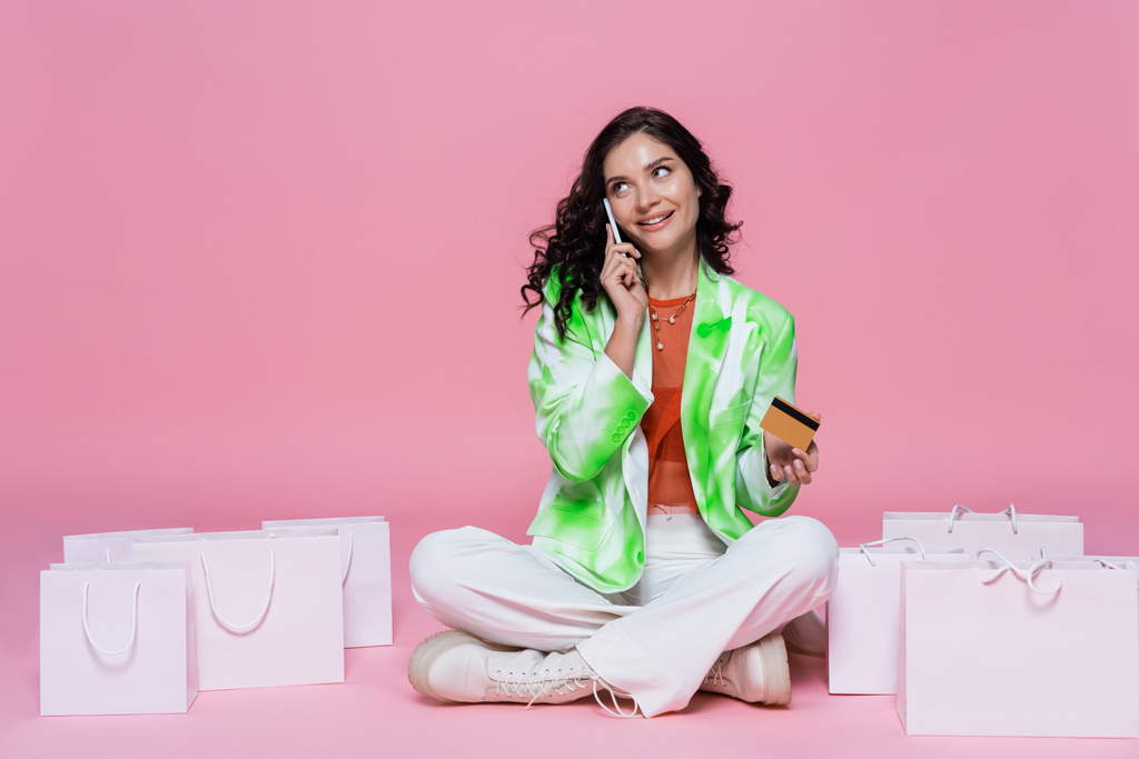 happy woman in blazer talking on smartphone while holding credit card near shopping bags on pink  - Photo, Image
