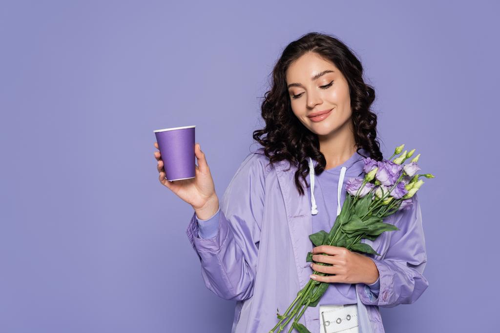 šťastná mladá žena v pláštěnce drží kytice květin a papírový pohár izolované na fialové - Fotografie, Obrázek