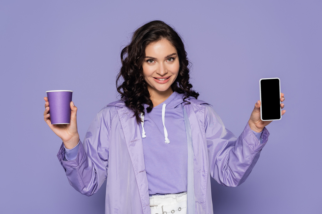 Yağmurluk giymiş mutlu genç bir kadın elinde kağıt bardak ve mor ekranlı akıllı telefon tutuyor. - Fotoğraf, Görsel