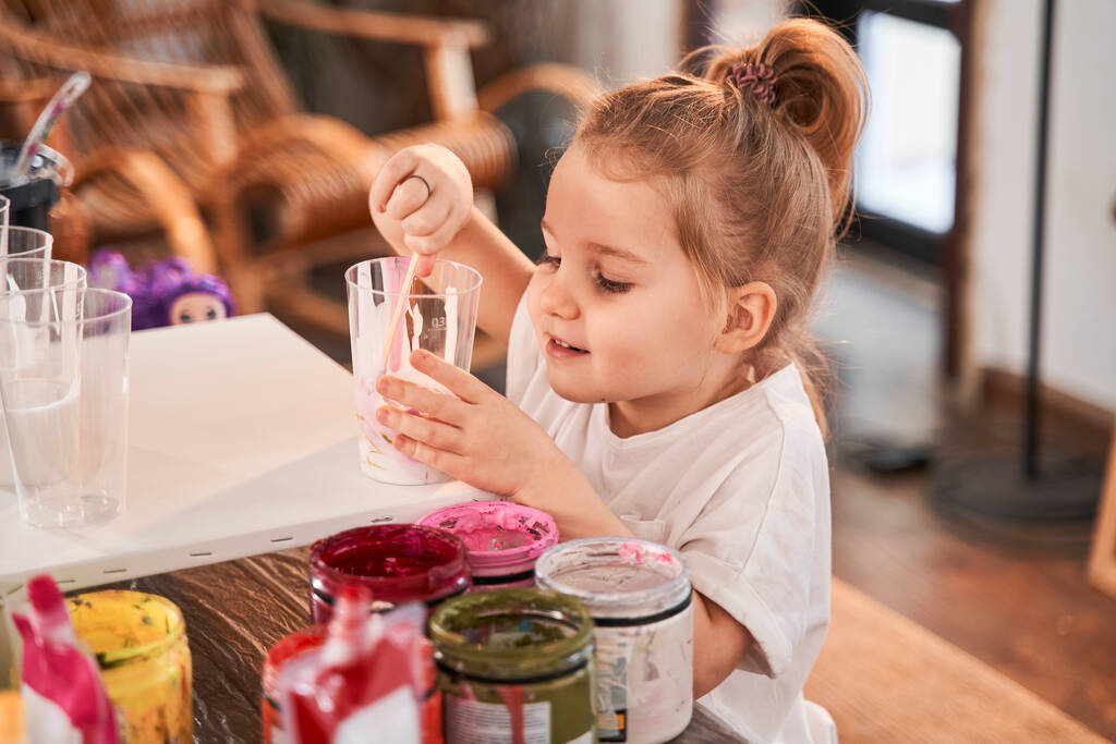 Девушка, стоящая за столом и держащая стекло красками, делая рисунки в художественной школе - Фото, изображение