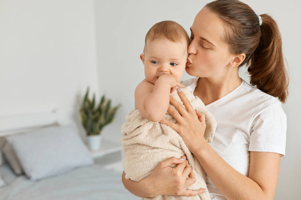 Indoor shot of young adult attractive woman holding daughter wrapped in towel after bathroom, mother besos her lovely baby girl, express love and gentle. - Foto, imagen