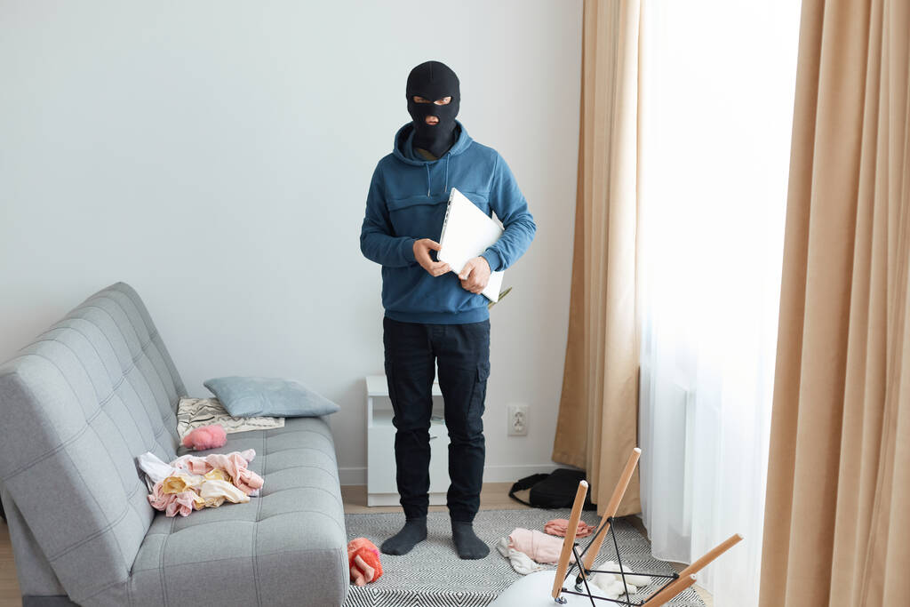 Человек крадет ноутбук в чужом доме, убегает, будучи преследуемым, носит маску грабителя и синюю толстовку, стоит рядом с диваном и окном, опасное ограбление ломается. - Фото, изображение