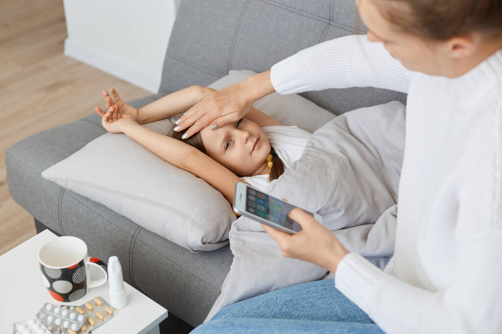 Πορτρέτο της γυναίκας που κάθεται στον καναπέ κοντά σε άρρωστη κόρη, κρατώντας το χέρι στο μέτωπό της και ποζάροντας με έξυπνο τηλέφωνο στα χέρια, καλώντας το γιατρό να ρωτήσει για τη θεραπεία. - Φωτογραφία, εικόνα