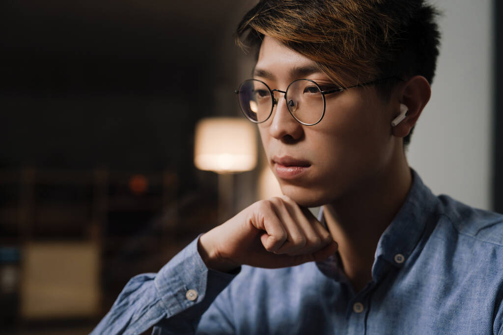 Focalisé asiatique guy dans lunettes à l'aide d'écouteurs sans fil tout en travaillant au bureau - Photo, image