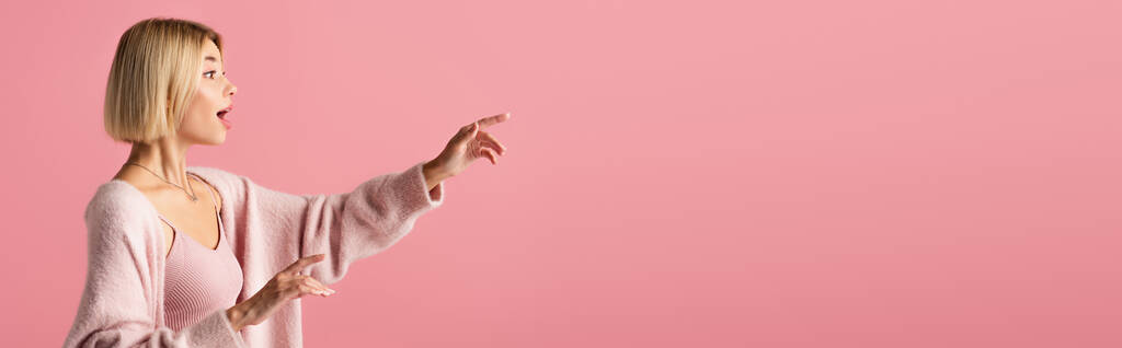side view of shocked young woman in soft cardigan looking away and gesturing isolated on pink, banner - Photo, Image
