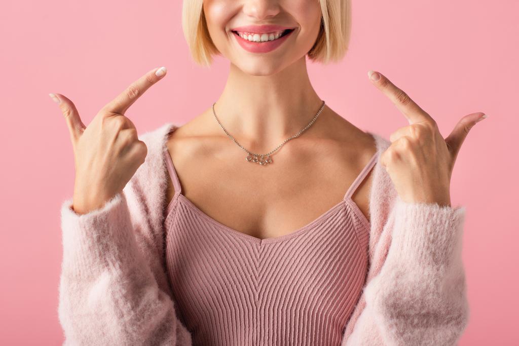 bijgesneden uitzicht van gelukkige jonge vrouw wijzen met vingers naar witte tanden geïsoleerd op roze  - Foto, afbeelding