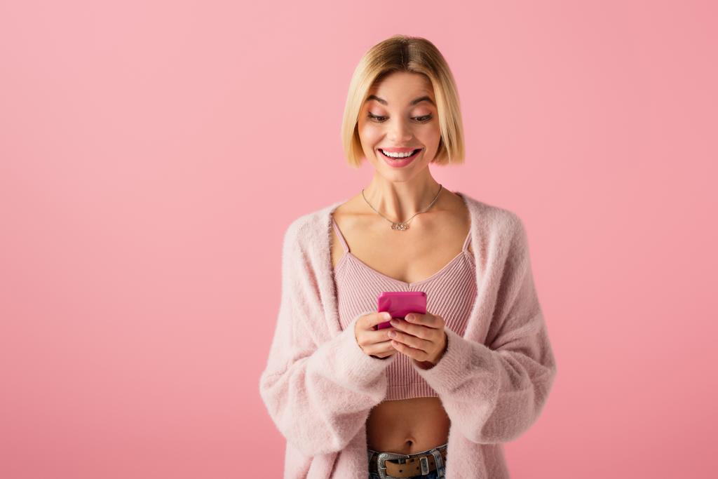 счастливая молодая женщина в мягком кардигане с помощью смартфона изолированы на розовый  - Фото, изображение